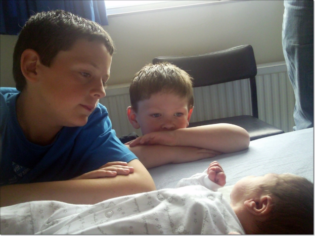 Ein Baby liegt auf dem Boden mit zwei Jungen links daneben, die das Baby anschauen, und einem Stuhl neben ihnen; ein Bein einer Person ist oben rechts sichtbar, mit einer Wand und einem blauen Vorhang im Hintergrund.