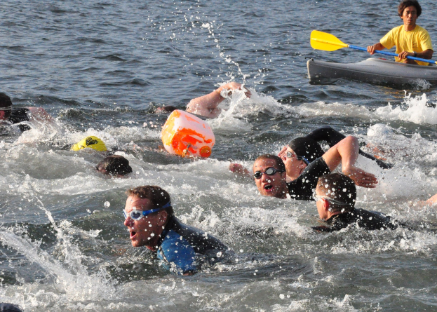 Gruppe von Menschen, die im Wasser schwimmen, mit einem Mann in einem Kajak, der ein Paddel hält, an einem Triathlon teilnimmt.
