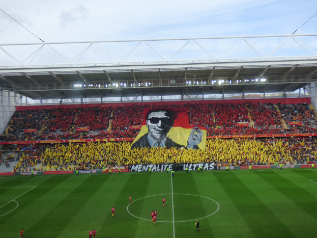 Ein Fußballstadion voller Menschen auf dem Feld und in den Rängen, mit einem zentralen Banner mit einem Porträt und Text, unter einem sichtbaren Himmel, Heimat von Watford Football Club.