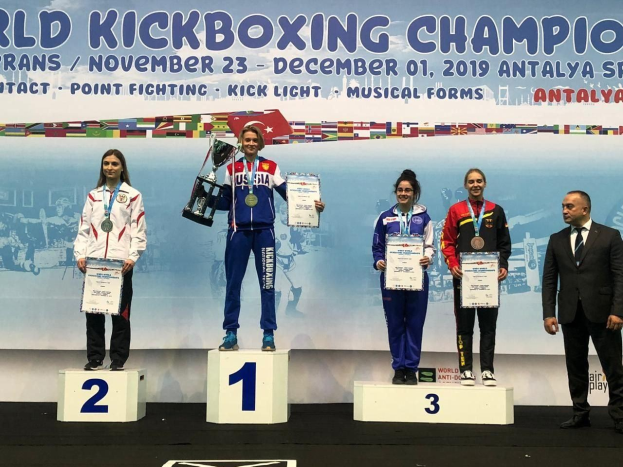 Fünf Personen mit Auszeichnungen und Urkunden auf einem Podest mit einem 'World Kickboxing Championship Antalya 2019'-Plakat im Hintergrund.