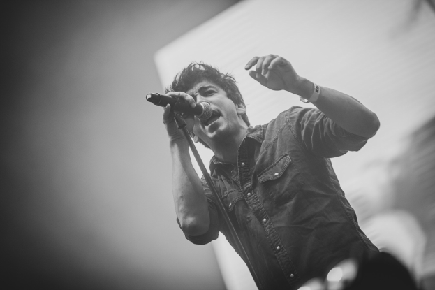 Schwarz-weiß-Foto eines Mannes, der in ein Mikrofon auf der Bühne singt, mit einem unscharfen Gesicht im Hintergrund.