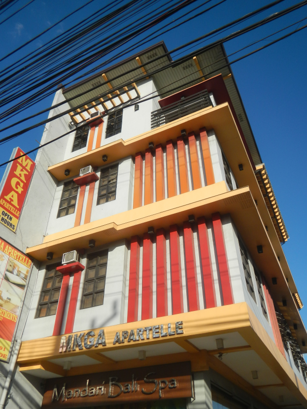 Ein Geb├Ąude mit Fenstern, Klimaanlagen, einem Schild mit der Aufschrift "Manga Apartments in Manila, Philippines" und Stromkabel unter einem bew├Âlktem Himmel.