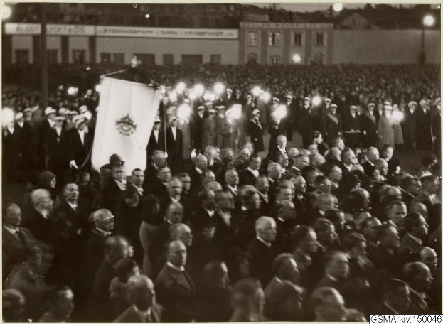 Schwarzes und weißes Foto einer großen Menge vor einem Gebäude, mit einer Person, die eine Fahne hält, Pfosten und Lichter im Hintergrund und einem Text am unteren Rand, der lautet: "Gustav Klimts Rede an der Berliner Mauer."