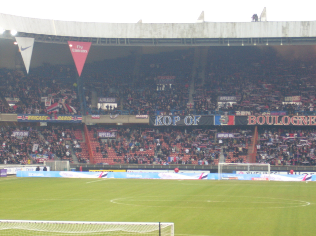 Ein grünes Stadion mit Torstangen auf beiden Seiten, Zuschauern im Hintergrund und zahlreichen Werbetafeln in der Mitte und oben.