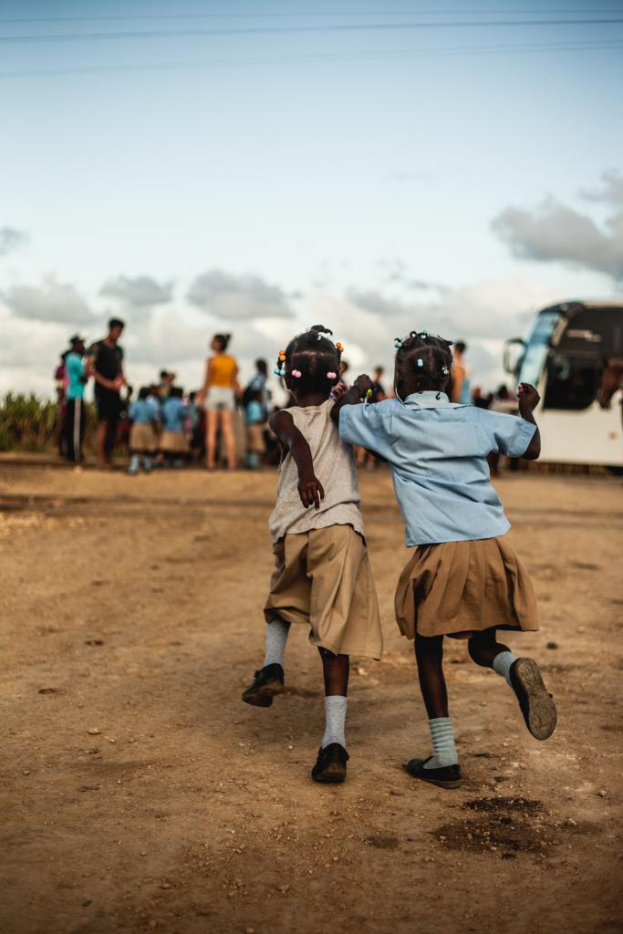 Zwei Kinder in Kleidern und Schuhen laufen, mit einem Fahrzeug auf der rechten Seite und einem Himmel darüber.