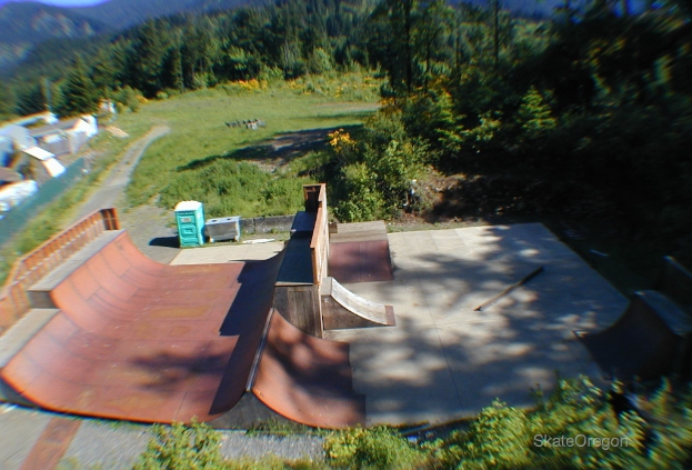Eine Skateanlage mit Boxen auf der linken Seite, Bäumen, Gras, Pflanzen und Blumen im Hintergrund und Text unten rechts.