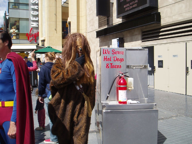 Eine Gruppe von Menschen, einige in Kostümen, versammelte sich vor Gebäuden mit Schildern, einer Box mit Schrift und einem Feuerlöscher im Hintergrund.