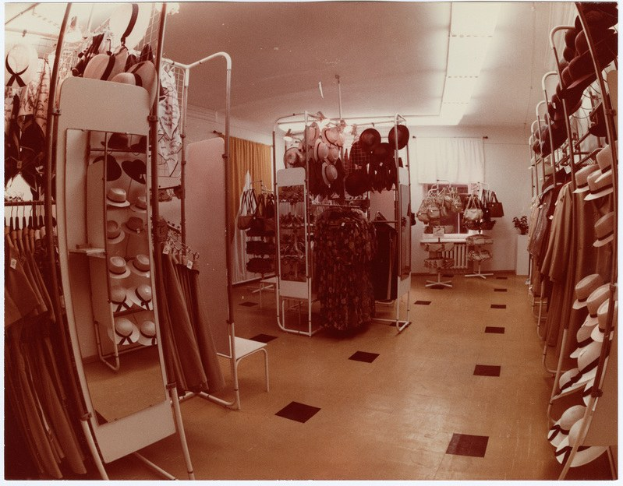 Altes Schwarz-Weiß-Foto eines kleinen Kleiderladens mit Kleiderstangen, Hutständern, einem Spiegel, einem Stuhl, einem Tisch mit diversen Gegenständen, gemusterten Vorhängen und Deckenleuchten.