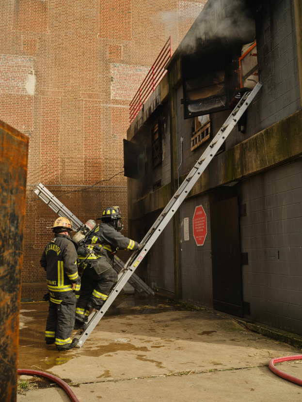 Feuerwehrleute mit Helmen klettern an einer Leiter zu einem rauchgefüllten Gebäude, während ein metallisches Objekt und Bodenschläuche sichtbar sind.