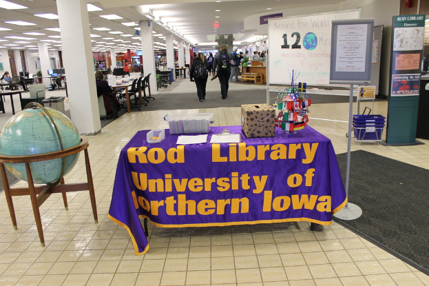 Eine Ausstellung mit einem Tisch, auf dem verschiedene Landesflaggen, eine Box, Papiere und ein Banner liegen, eine Kugel links daneben, ein Anschlagbrett rechts daneben mit einigen Menschen in der Nähe und mehreren Tischen und Stühlen im Hintergrund.