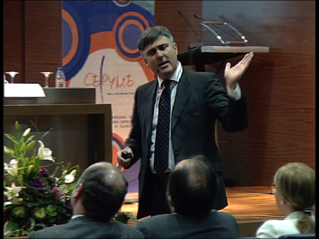 Drei Personen sitzen in der ersten Reihe eines Konferenzsaals, ein Mann in einem schwarzen Blazer steht und spricht in ein Mikrofon, ein Blumenarrangement und ein Tisch mit einem Buch und einer Wasserflasche in der Nähe, ein Podium mit Mikrofonen dahinter und eine farbenfrohe Tafel mit Text im Hintergrund.