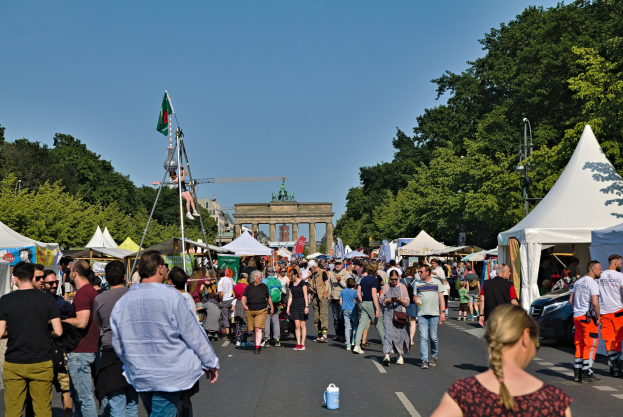 Eine Menschenmenge, die eine Straße mit Zelten, Fahrzeugen und Bäumen entlanggeht, mit einem Bogen und einem klaren blauen Himmel im Hintergrund und Fahnenmästen auf der linken Seite.