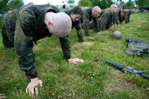 Eine Gruppe von Männern in Militäruniformen führt Liegestütze auf dem Gras durch, umgeben von verstreuter Ausrüstung, mit Bäumen, Fahrzeugen und einem klaren blauen Himmel im Hintergrund.