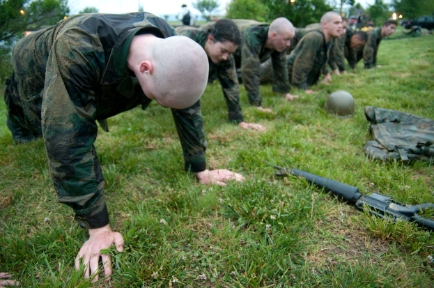 Eine Gruppe von Männern in Militäruniformen führt Liegestütze auf dem Gras durch, umgeben von verstreuter Ausrüstung, mit Bäumen, Fahrzeugen und einem klaren blauen Himmel im Hintergrund.