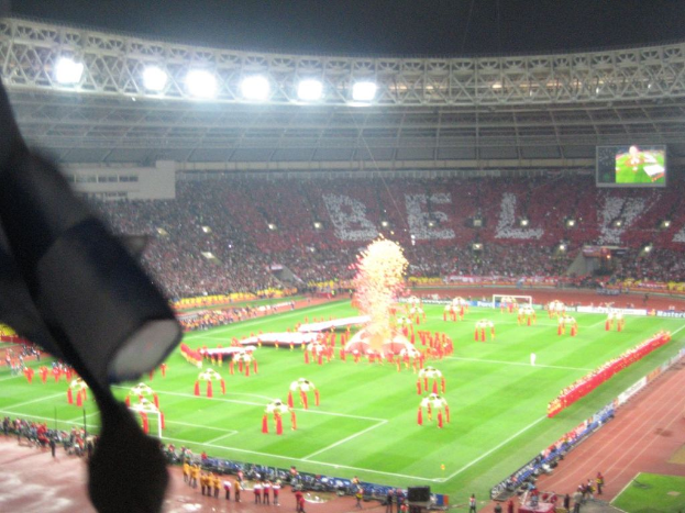 Ein Stadion mit Flutlicht, Eisengittern, Zuschauern, stehenden Personen, Werbetafeln, Anzeigetafeln, Ãberwachungskameras, Flaggen und DÃ©corations.