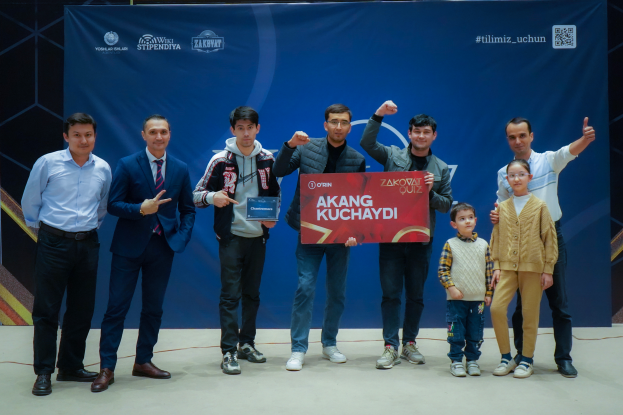 Eine Gruppe von Menschen auf einer Bühne, die eine Tafel mit dem Text "Akang Kuchaydi - Zakovat Quiz" halten, mit einem Banner, Lichtern und anderen Gegenständen im Hintergrund.