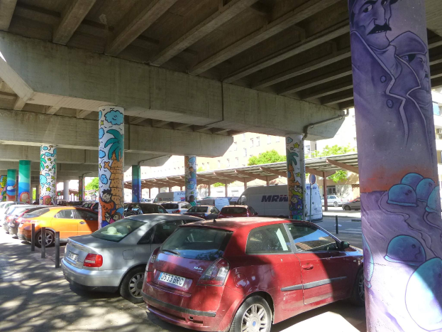 Ein Parkplatz voller parkender Autos unter einer Brücke mit Graffiti an den Säulen und Gebäuden und Bäumen im Hintergrund, inklusive eines Lieferwagens.