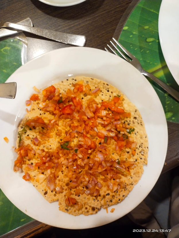Ein weißer Teller mit einem Fladenbrot, das mit Tomaten, Zwiebeln und Paprika belegt ist und von Besteck auf einem Tisch begleitet wird.