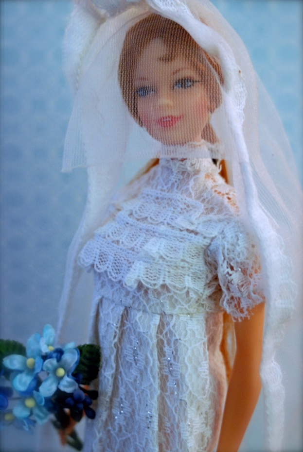 Eine Puppe in einem Kleid, Schleier und mit einem Strauß, vor einem Objekt.