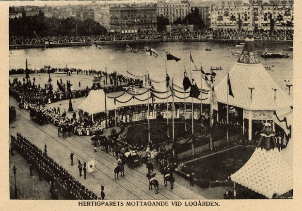 Ein altes Schwarz-Weiß-Foto einer Menge, die sich um einen Karneval mit Zelten, Fahnen, Masten und Pferden im Vordergrund und Gebäuden, Bäumen und Wasser im Hintergrund versammelt hat, mit Text am unteren Bildrand.