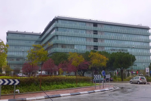 Großes modernes Gebäude mit vielen Fenstern, der Hauptsitz der Europäischen Zentralbank, umgeben von Grünflächen, Straßeninfrastruktur und Fahrzeugen unter einem bewölkten Himmel.