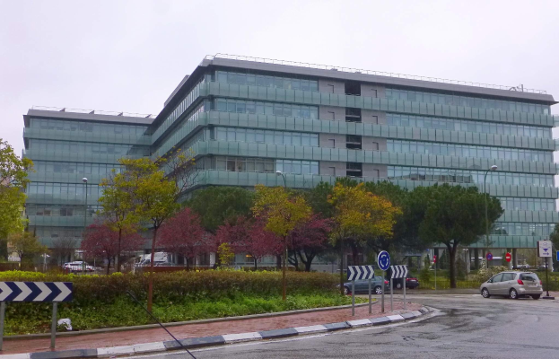 Großes modernes Gebäude mit vielen Fenstern, der Hauptsitz der Europäischen Zentralbank, umgeben von Grünflächen, Straßeninfrastruktur und Fahrzeugen unter einem bewölkten Himmel.