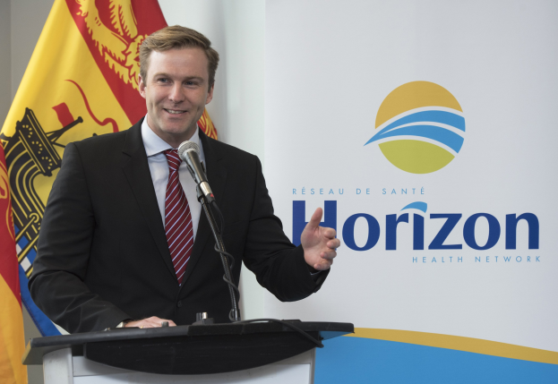 Ein l├Ąchelnder Mann in einer schwarzen Jacke, weißem Hemd und roter Krawatte steht an einem Rednerpult mit einem Mikrofon und einem Banner im Hintergrund.