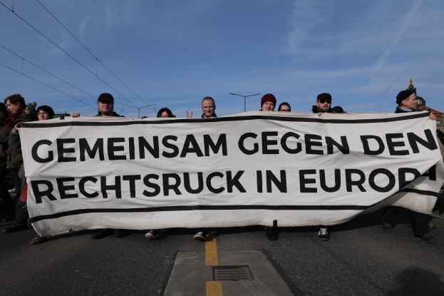 Eine Gruppe von Menschen steht auf einer Straße und hält ein Banner mit der Aufschrift "Gemeinsam Gegen den Rechtsruck in Europa" in den Händen mit Straßenlaternen, Strommasten, Stromkabeln, Gebäuden und einem bewölkten Himmel im Hintergrund.