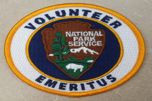 Ein rundes Patch mit einem blauen Rand, weißem Hintergrund und fetter schwarzer Schrift, die 'Volunteer National Park Service Emeritus' in der Mitte eines gelben Kreises liest.