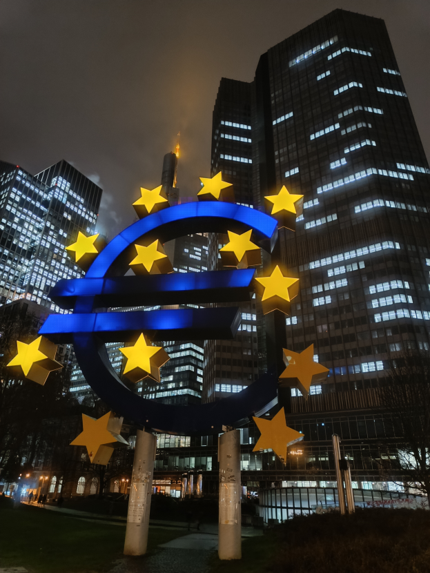 Das Euro-Zeichen steht prominent vor der beleuchteten Zentrale der Europäischen Zentralbank in Frankfurt, Deutschland, umgeben von Bäumen, Pflanzen und Gras, mit dem Himmel im Hintergrund.