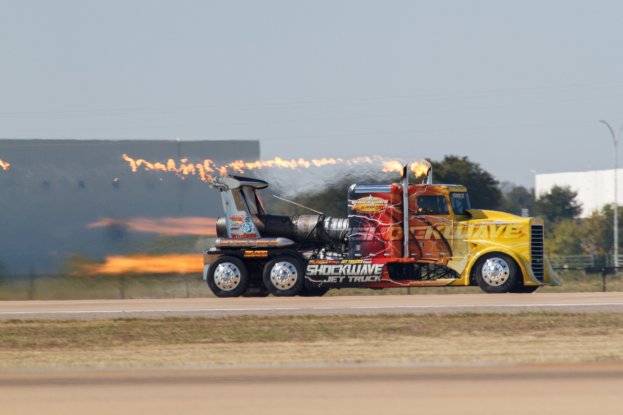 Flammen schlagen aus dem Heck eines Lastwagens, der auf einer Straße fährt, die von Gras, einem Zaun, Bäumen, einem Gebäude, einem Strommast, Drähten und einem bewölkten Himmel umgeben ist, mit einem sichtbaren Reifen und Rad.