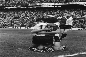 Drei Fußballspieler umarmen sich auf dem Boden und feiern ein Tor vor einem jubelnden Publikum in einer schwarz-weißen Stadionkulisse.