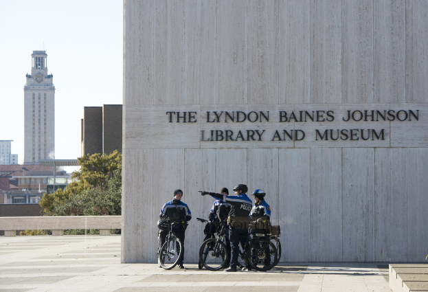 Eine Gruppe von Polizisten in blauen Uniformen und Helmen steht mit Fahrrädern vor dem Lyndon Baines Johnson Library and Museum, mit einer texttragenden Wand, Treppen, Bäumen, Gebäuden und einem klaren blauen Himmel im Hintergrund.