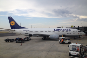 Ein Lufthansa Airbus A320-200 auf dem Frankfurt Airport geparkt, mit verschiedenen Bodenfahrzeugen, Wagen, Verkehrskegeln, Polen und Texttafeln im Hintergrund, unter einem bewölkten Himmel.