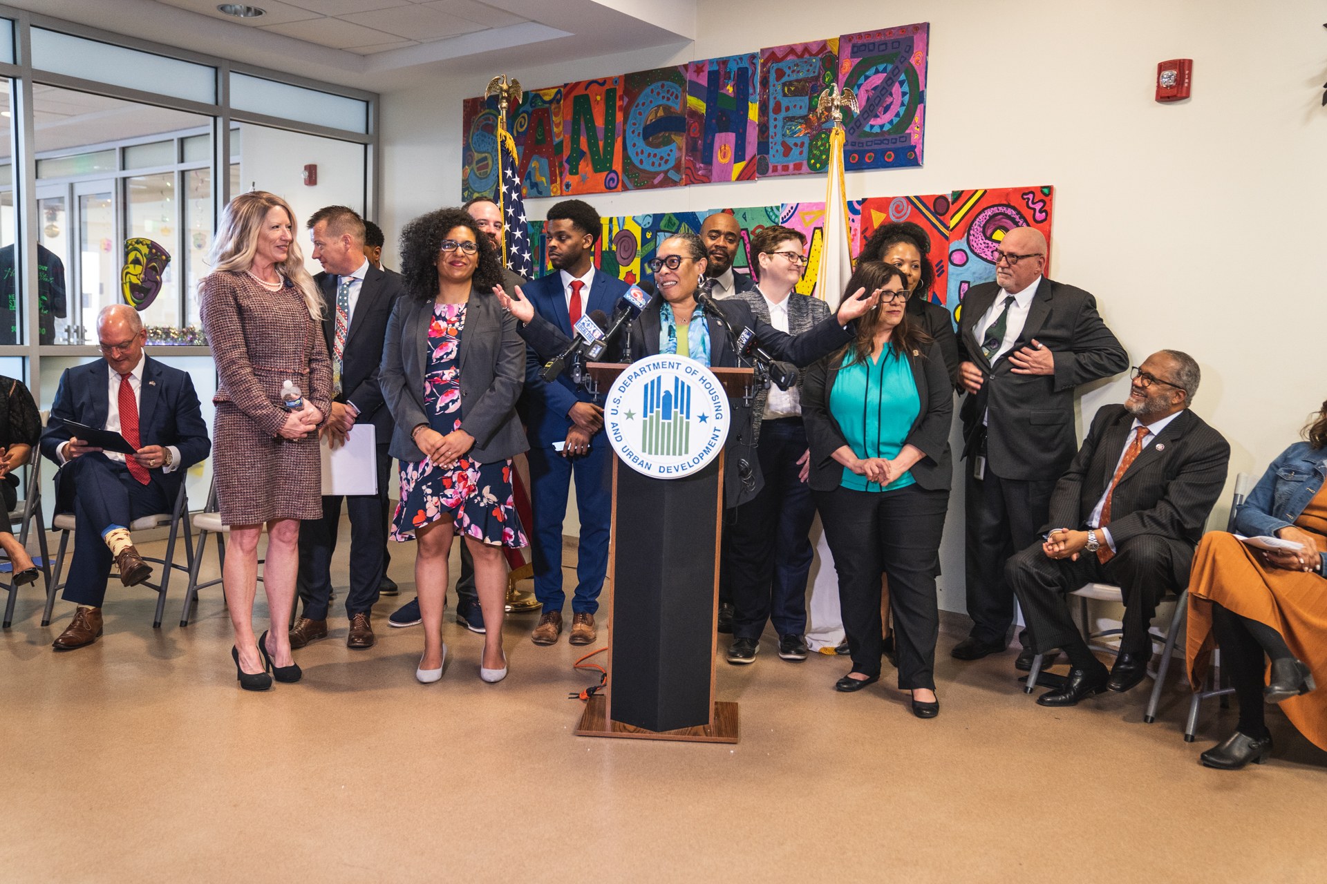 Gruppe von Menschen vor einem Podium mit einem Mikrofon und einer Tafel mit der Aufschrift "Sancho Stadtrat tritt dem Stadtrat bei", mit sitzenden und stehenden Zuschauern, Flaggen, einem Feuerlöscher und Deckenleuchten im Hintergrund.