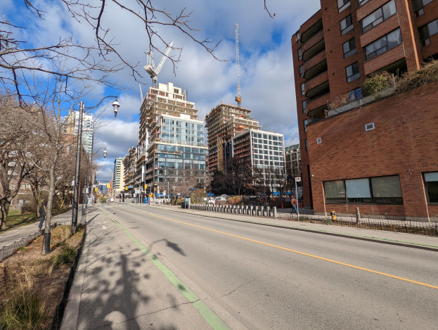 Eine Stadtstraße mit hohen Gebäuden, Straßenlaternen, Stromleitungen, Fahrzeugen, Fußgängern, Geländern, Bäumen und einer bewölkten Himmel.