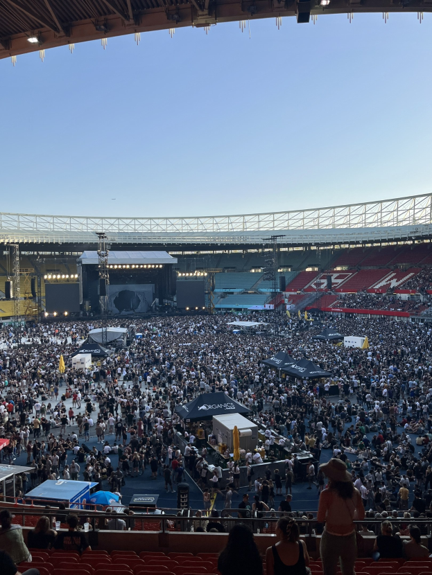 Eine große Menschenmenge sitzt und steht in einem Stadion mit Zelten, Geländern und Scheinwerfern, was auf ein Konzert in der O2 Arena in London hindeutet.