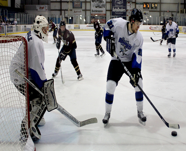 Eine Gruppe junger M├Ąnner, die auf einem Eisplatz Eishockey spielen, tragen Helme und halten Hockeyschl├Ąger, mit einem Torpfosten links und Bannern im Hintergrund.