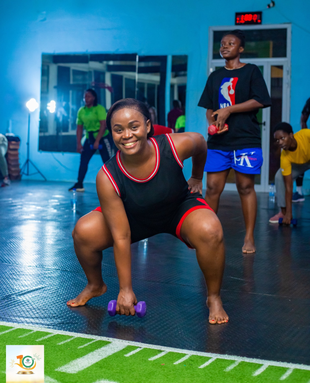 Eine Frau in Kniebeugeposition mit Hanteln in einer Fitnessstudio-Umgebung mit Equipment und Branding im Hintergrund.