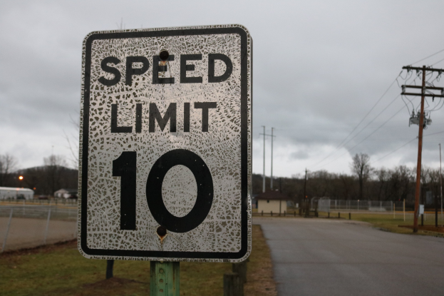 Tempolimitschild auf der Seite einer Straße mit Gras, Strommasten, einem Zaun, Häusern, Bäumen und einem bewölkten Himmel im Hintergrund.