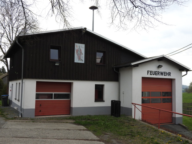 Rote und weiße Feuerwache mit Straße, Gras, gelben Blumen und Bäumen im Vordergrund und Himmel im Hintergrund.