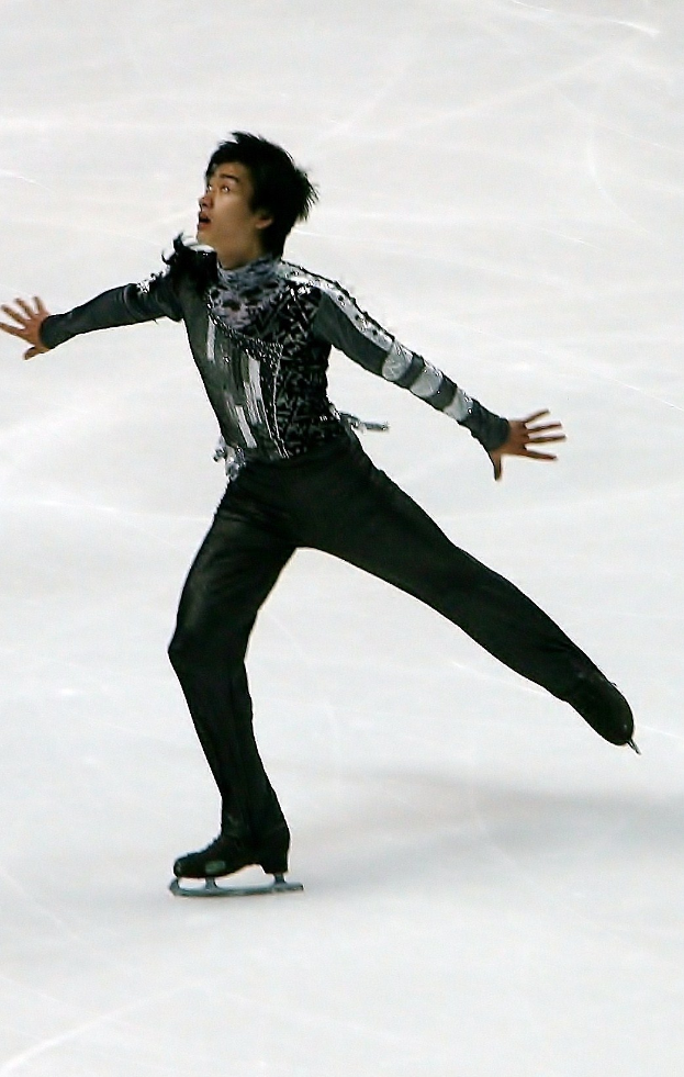 Männlicher Eiskunstläufer gleitet über eine Eisfläche in einem schwarzen Kleid und Schlittschuhen, führt eine Choreographie mit synchronen Arm- und Beinbewegungen aus.