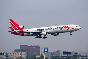 Ein Martinair Cargo Boeing 777-300ER startet am Frankfurter Flughafen, mit Gebäuden, Bäumen, Pfählen und Werbetafeln im Hintergrund und dem Himmel oben.