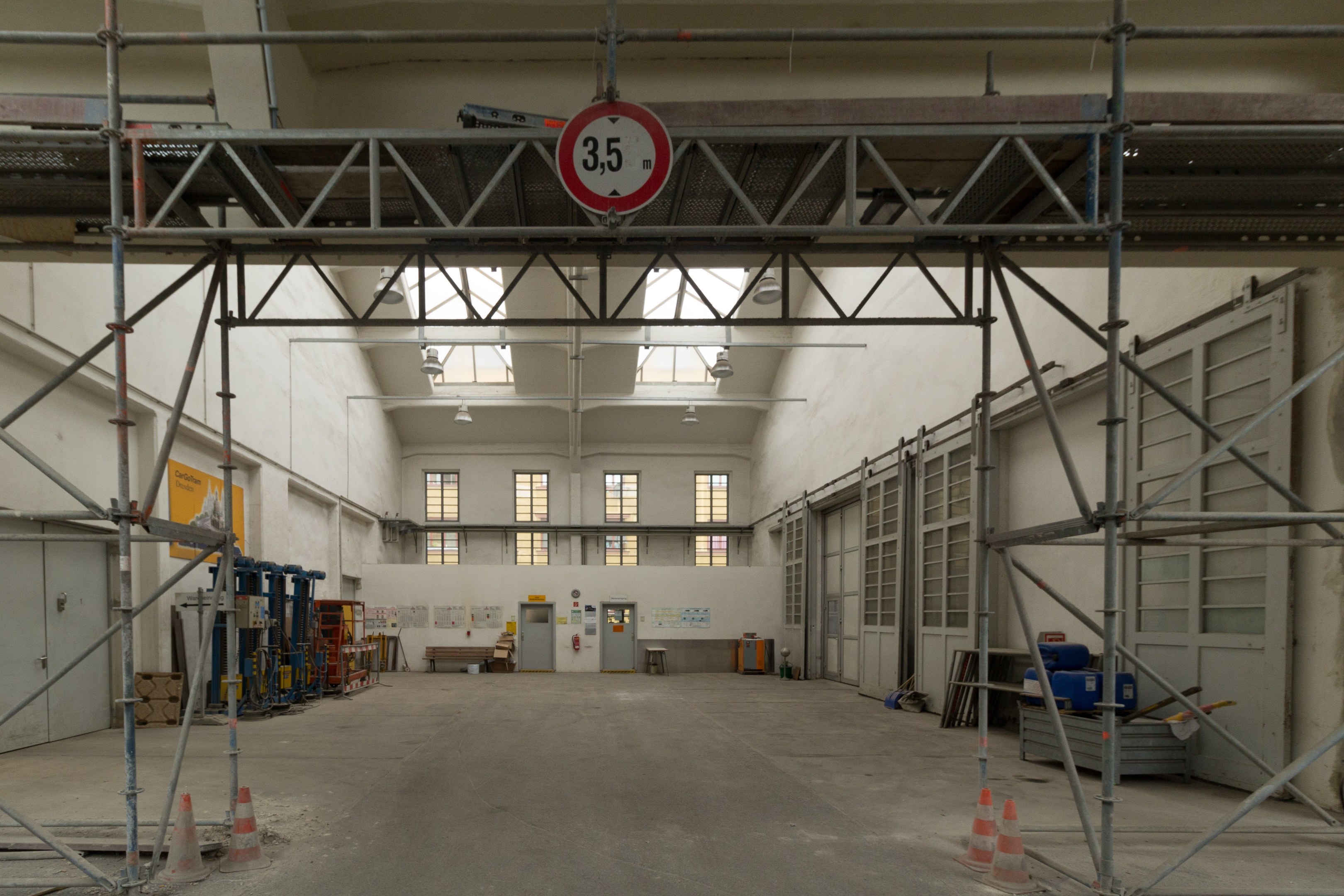Großes Lagerinnere mit ausgedehnten Gerüsten, Verkehrskegeln, Metallstangen, einem Schild, Maschinen, einer Tür, Fenstern, Wänden, Deckenlampen und Dach.