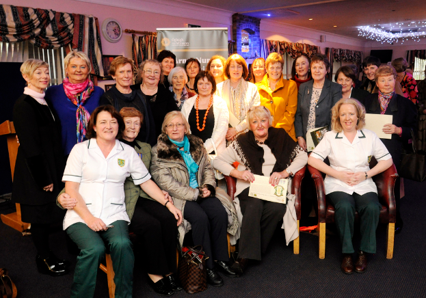 Fünf Frauen im Vordergrund, viele Frauen in der Mitte, mit Plakaten, einem Vorhang, einer Uhr, Fenstern, Lampen, einem Boden und einer Wand im Hintergrund.