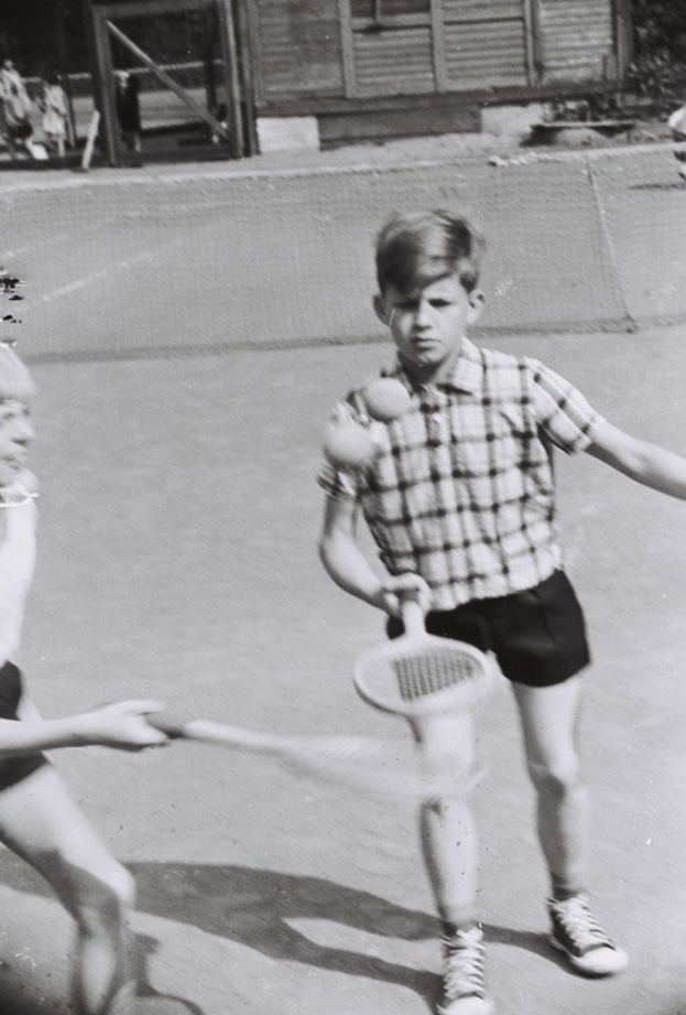 Zwei junge Jungs spielen Tennis auf einem schwarz-weißen Court, jeder hält einen Schläger, mit einem Netz, Menschen, Bäumen und einem Gebäude im Hintergrund.