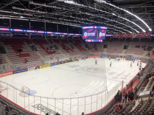 Eishockeyspiel in einer großen Arena zwischen den St. Louis Blues und Detroit Red Wings, mit Zuschauern auf den Sitzplätzen und in der Nähe der Eisfläche stehend, umgeben von einem Zaun, einem Bildschirm, Metallrahmen, Deckenlampen und einem Dach.