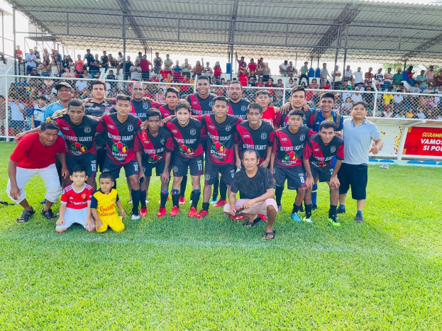 Gruppe von Männern auf einem Fußballfeld mit einem Siegesschild, das "Fotos Ugarte U-17 vs U-18" trägt, unter einem bewölkten Himmel.