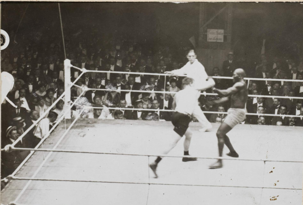 Zwei Boxer im Ring, umgeben von einer Menge, mit Texttafeln an der Wand hinter ihnen, die ihren Kampf feiern.