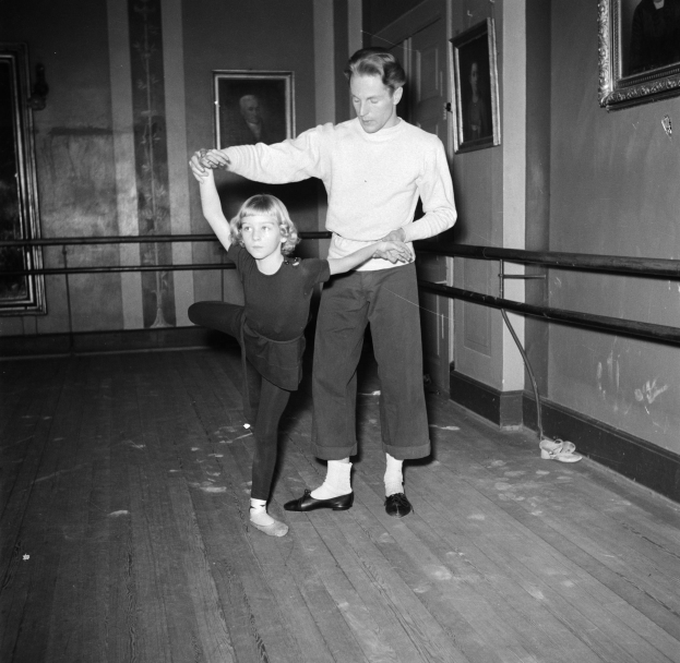 Ein Mann in einem weißen Hemd und schwarzen Hosen tanzt mit einem kleinen Mädchen in einem Tanzstudio, mit Fotorahmen an den Wänden und einem Geländer im Hintergrund, alles in Schwarz-Weiß dargestellt.